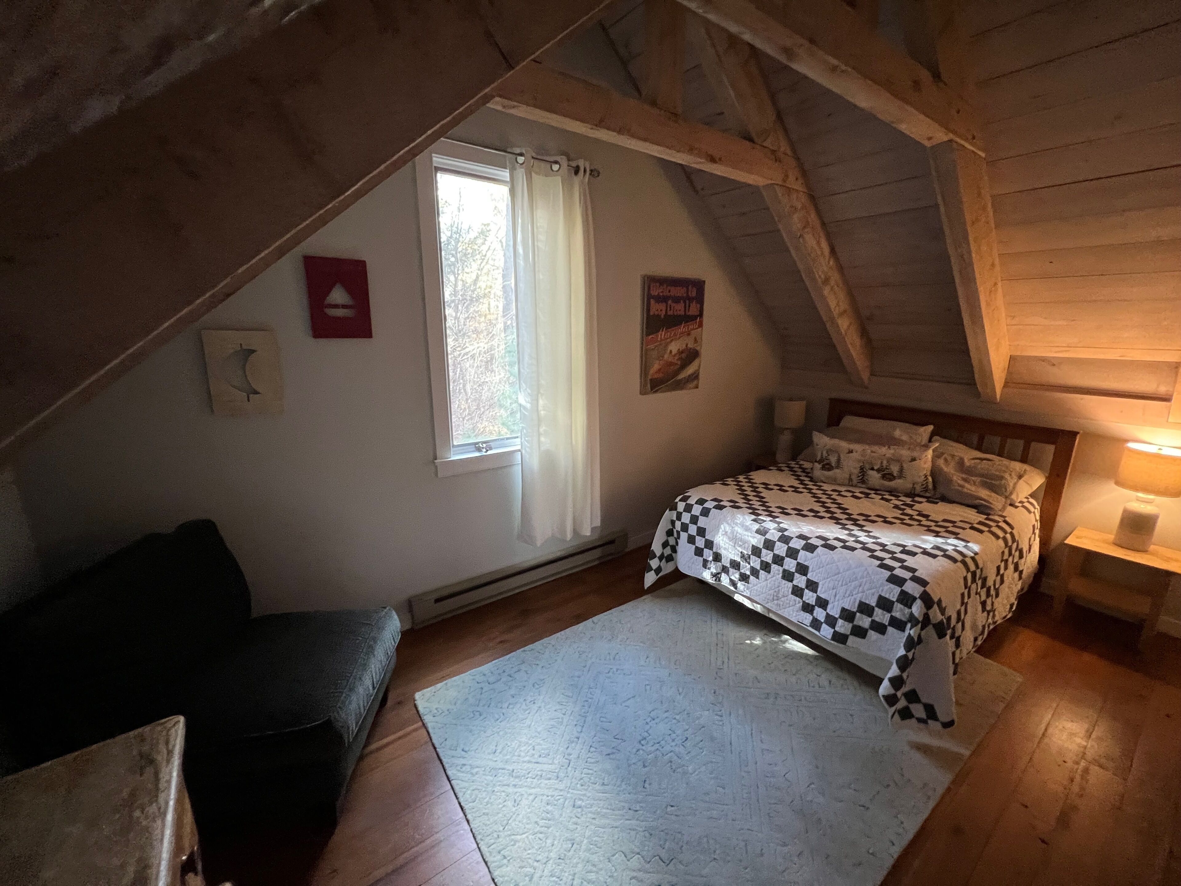 Un dormitorio acogedor en el ático, con colcha a cuadros, una ventana pequeña con cortinas y techo de madera.