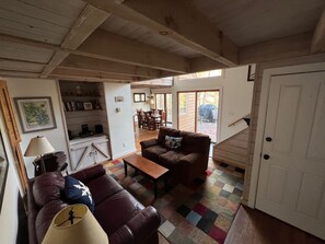 Una sala de estar acogedora con un sofá de cuero marrón, una mesa de centro de madera y un tapete colorido. Se ve un comedor con mesa y sillas a través de una puerta de vidrio.