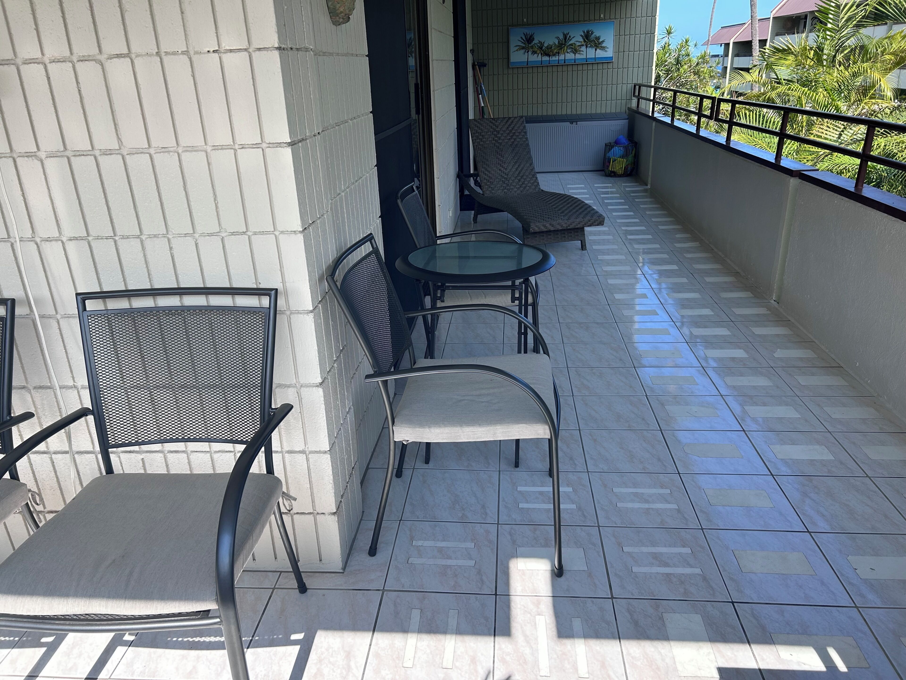 A balcony with tiled flooring, metal chairs, a glass table, and a view of palm trees and buildings.