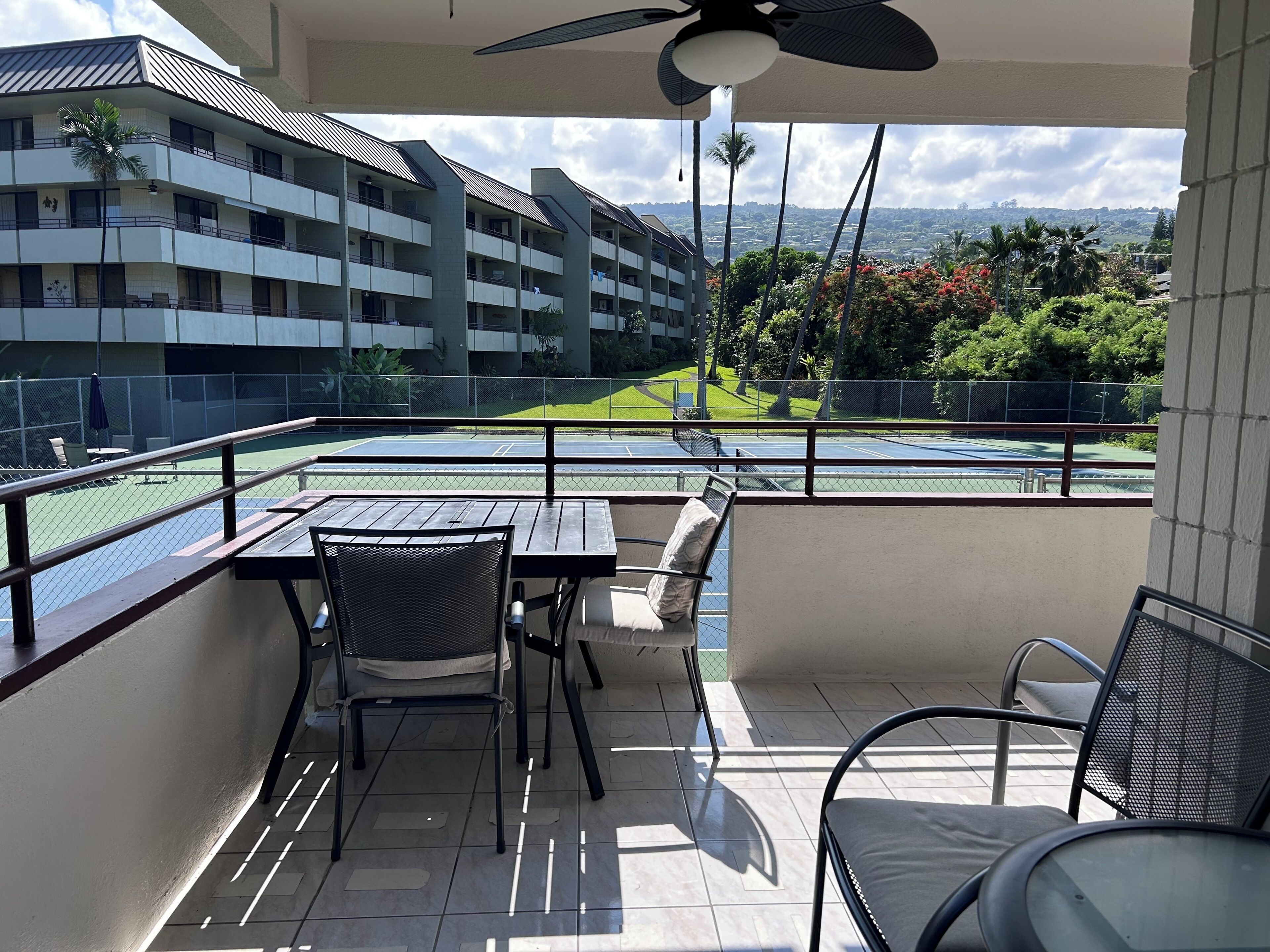 Ein Balkon mit Tisch und Stühlen, einem Deckenventilator und Blick auf einen Tennisplatz sowie Gebäude.