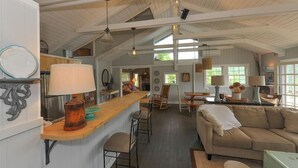 Un vaste espace de vie doté d’un haut plafond et de poutres apparentes, avec un coin salon central comprenant un canapé et des fauteuils, ainsi qu’un coin cuisine avec un bar et une table à manger.