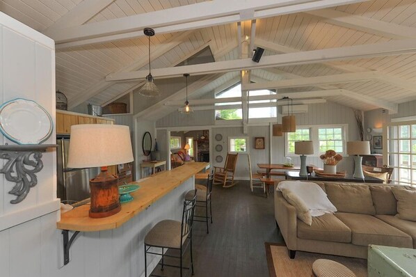A spacious living area with a high ceiling, wooden beams, a central seating area with a sofa and chairs, and a kitchen area with a bar counter and dining table.