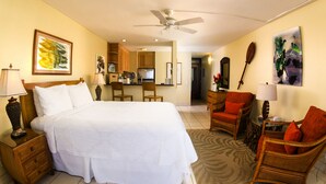 Habitación de hotel con una cama grande, un escritorio, dos sillas, un ventilador de techo y una cocina al fondo.