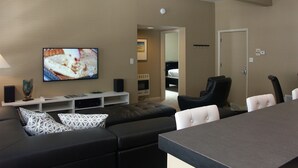 Ein modernes Wohnzimmer mit einer schwarzen Ledersofa, einem Wand-Fernseher, einem Esstisch mit Stühlen und einem weißen Lowboard mit Lautsprechern.