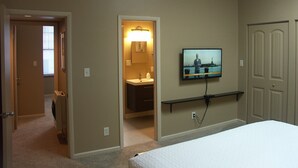 Ein Schlafzimmer mit einem Bett, einem an der Wand montierten Fernseher, einer Tür und einem Flur, der zu einem Badezimmer führt.