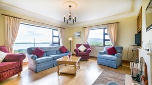 A spacious living room with a large window offering a scenic view, a chandelier, and a fireplace. - Self-Catering Holiday Home in Beautiful Ventry (Dingle)