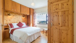 A bedroom with a wooden wardrobe, a bed with white bedding and red pillows, a small table, and a window with a view of the outside. - Self-Catering Holiday Home in Beautiful Ventry (Dingle)