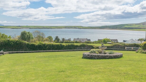 A grassy area with a stone fountain, a stone wall, and houses in the background near a body of water. - Self-Catering Holiday Home in Beautiful Ventry (Dingle)