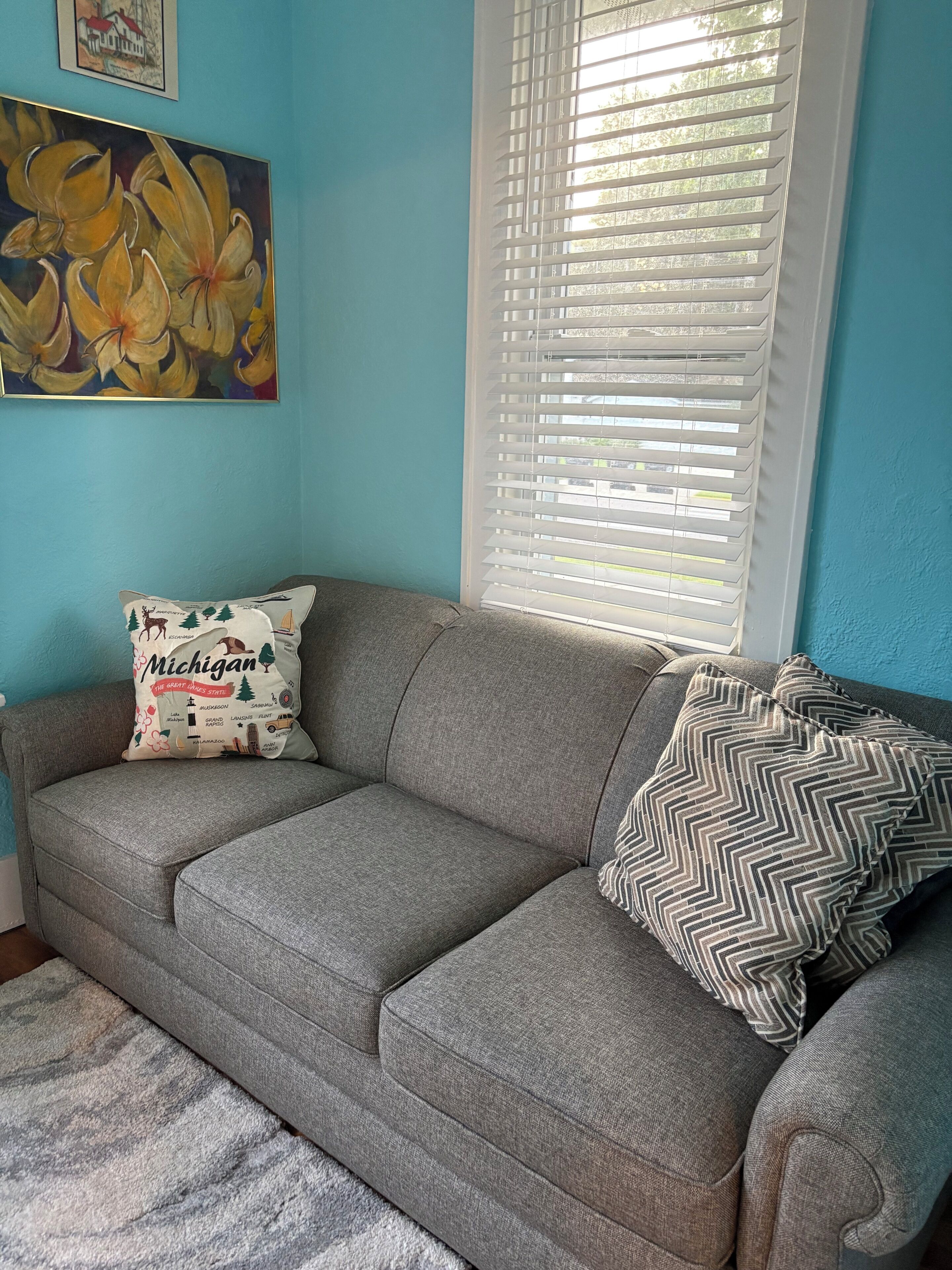 A gray sofa with two pillows, one with a Michigan-themed design, and a painting of yellow flowers on a blue wall.