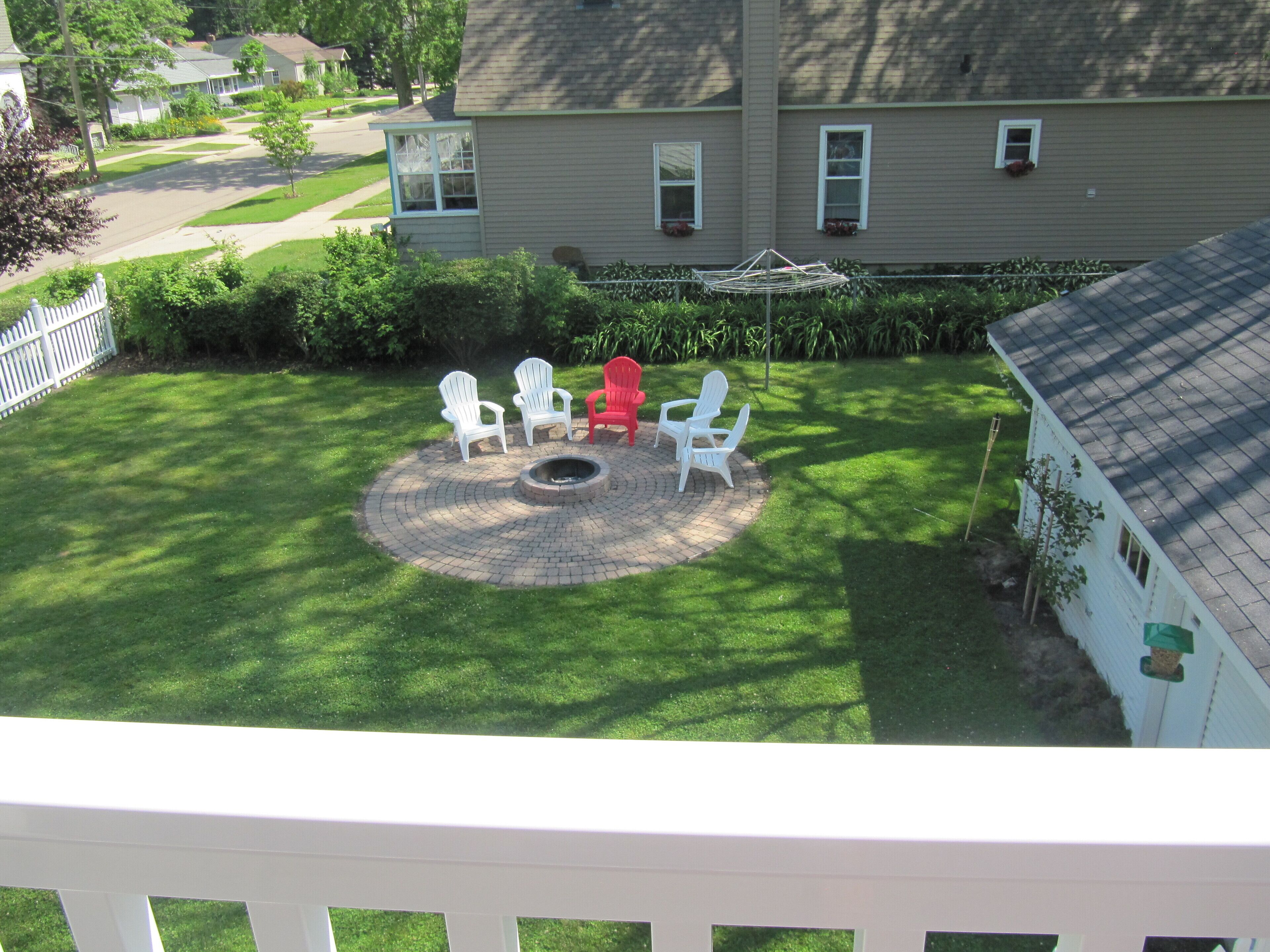 A backyard with a circular fire pit, four Adirondack chairs, and a red chair.