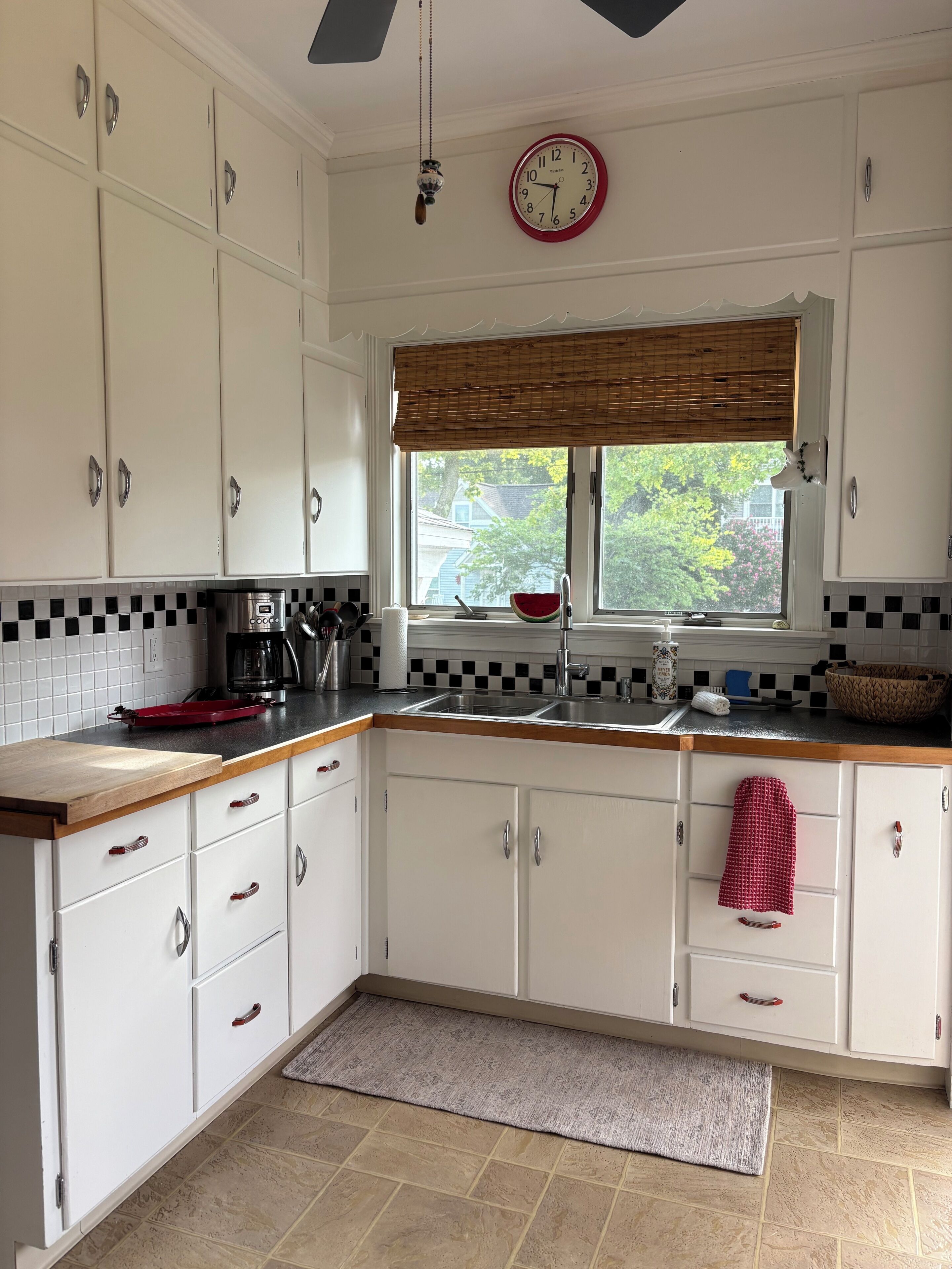 A kitchen with white cabinets, a wooden countertop, a sink, a clock, and a window with a view of greenery.