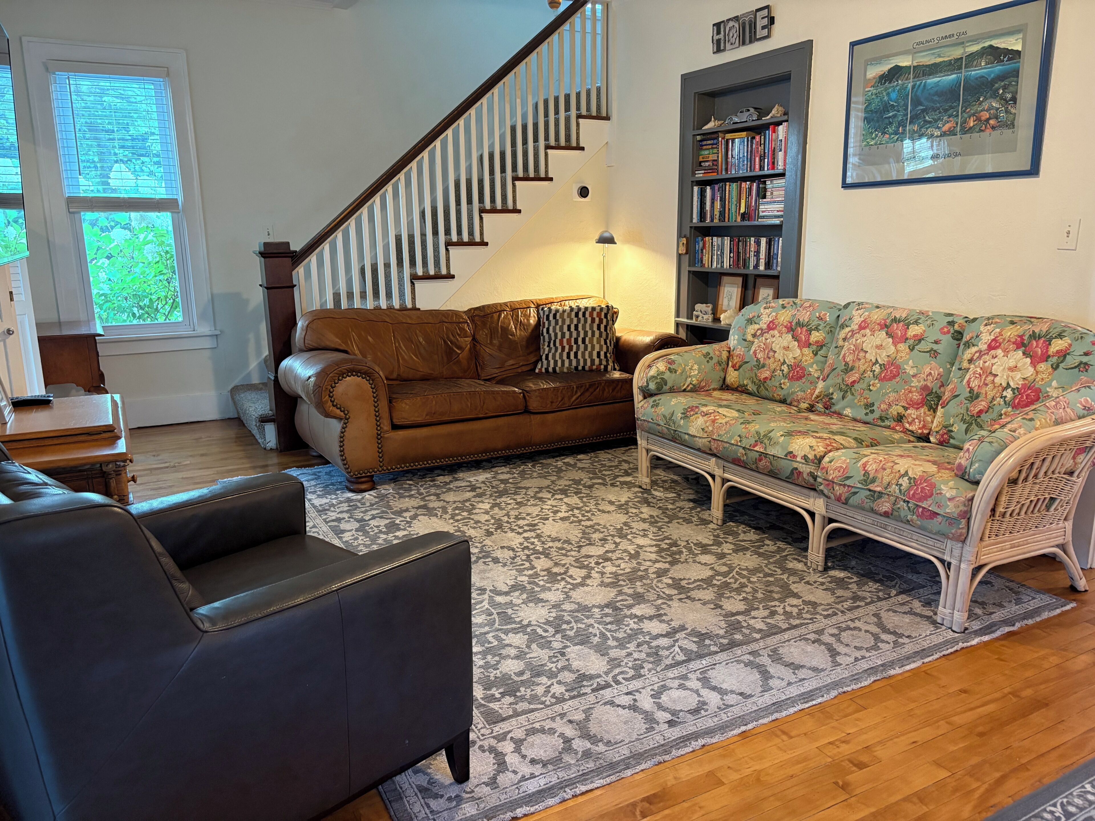 A living room with a brown leather sofa, a floral-patterned sofa, a wicker chair, a wooden coffee table, a bookshelf, and a staircase.