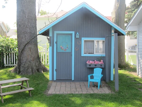 A small, single-story shed with a blue door and window, a bench, and a chair outside.
