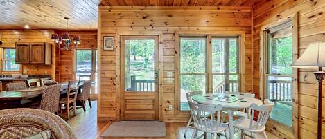 A wooden cabin interior with a dining area, a living room with a sofa, and large windows offering a view of the outdoors.