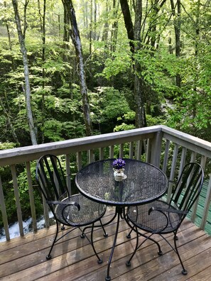 A wooden deck with a round metal table and two chairs, surrounded by lush greenery.