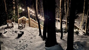 Un espacio exterior cubierto de nieve con bancos de madera y una valla de madera, iluminado por luces colgantes.