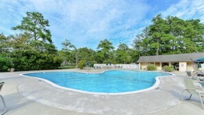 En swimmingpool med en buet kant, omgivet af en beton terrasse. Loungestole og en bygning med et skråt tag i baggrunden.