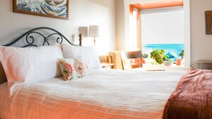 Ein ordentlich bezogenes Bett mit weißer und orangefarbener Bettwäsche, ein gerahmtes Kunstwerk darüber und ein Fenster mit Blick aufs Meer.