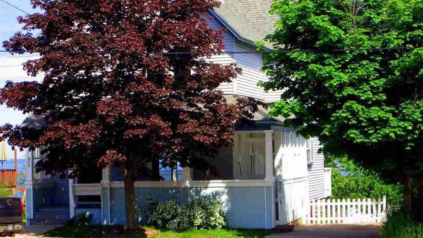 Ein zweistöckiges Haus mit Veranda, ein Baum mit roten Blättern und ein weißer Zierzaun.