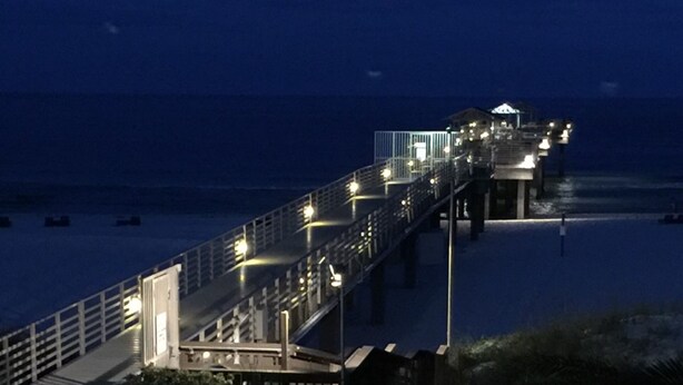 A beachside walkway leading to a building with a pool area featuring sun loungers and a potted plant.