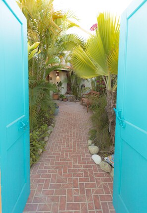 Une porte turquoise qui donne sur un chemin de briques, bordé d’une végétation luxuriante et de plantes en pot.