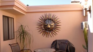 An outdoor patio with two metal chairs, a glass table, a decorative sun-like wall art, a cactus, and a covered grill. - BEAUTIFUL SUNRISES.  THREE MASTER SUITES (Palm Desert)