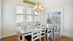 A dining area with a glass-top table and white chairs, a chandelier, and a sign reading "BEACH HOUSE". - 5543-C-Dreams - Nags Head Ocean View Home | Private Pool (Nags Head)