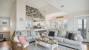 A spacious living room with a grey sectional sofa, a wicker chair, and a glass coffee table. A kitchen area is visible in the background with white cabinets and a sign reading "BEACH HOUSE". - 5543-C-Dreams - Nags Head Ocean View Home | Private Pool (Nags Head)