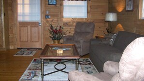A cozy log cabin interior with a wooden floor, a gray sofa, a matching armchair, a coffee table with a vase of flowers, and a window with blinds.