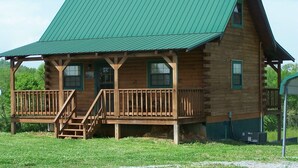 A wooden cabin with a green metal roof, a porch with stairs, and a small window on the side.