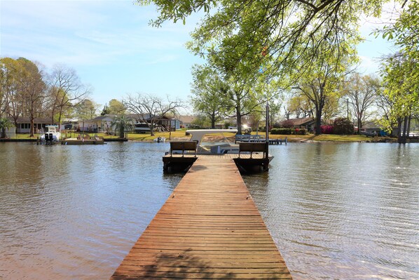 A wooden dock extending into a calm lake with benches on either end, surrounded by trees and houses in the background.