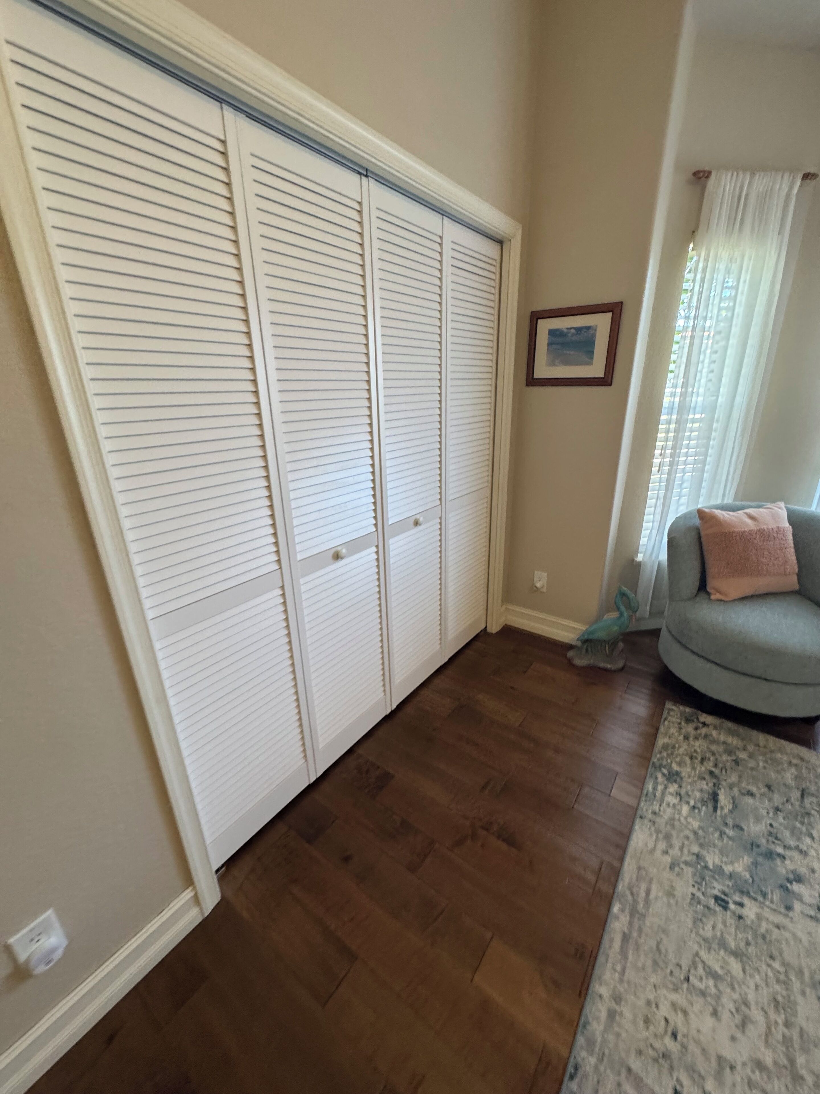 A room with a wooden floor, a grey sofa with a pink pillow, and a white closet with horizontal blinds.