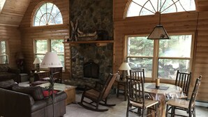 Interior acogedor de una cabaña con chimenea de piedra, mobiliario de madera y grandes ventanas con vistas al bosque.