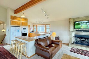 A modern kitchen with wooden cabinets, a central island with stools, a fireplace, and a television mounted above it.