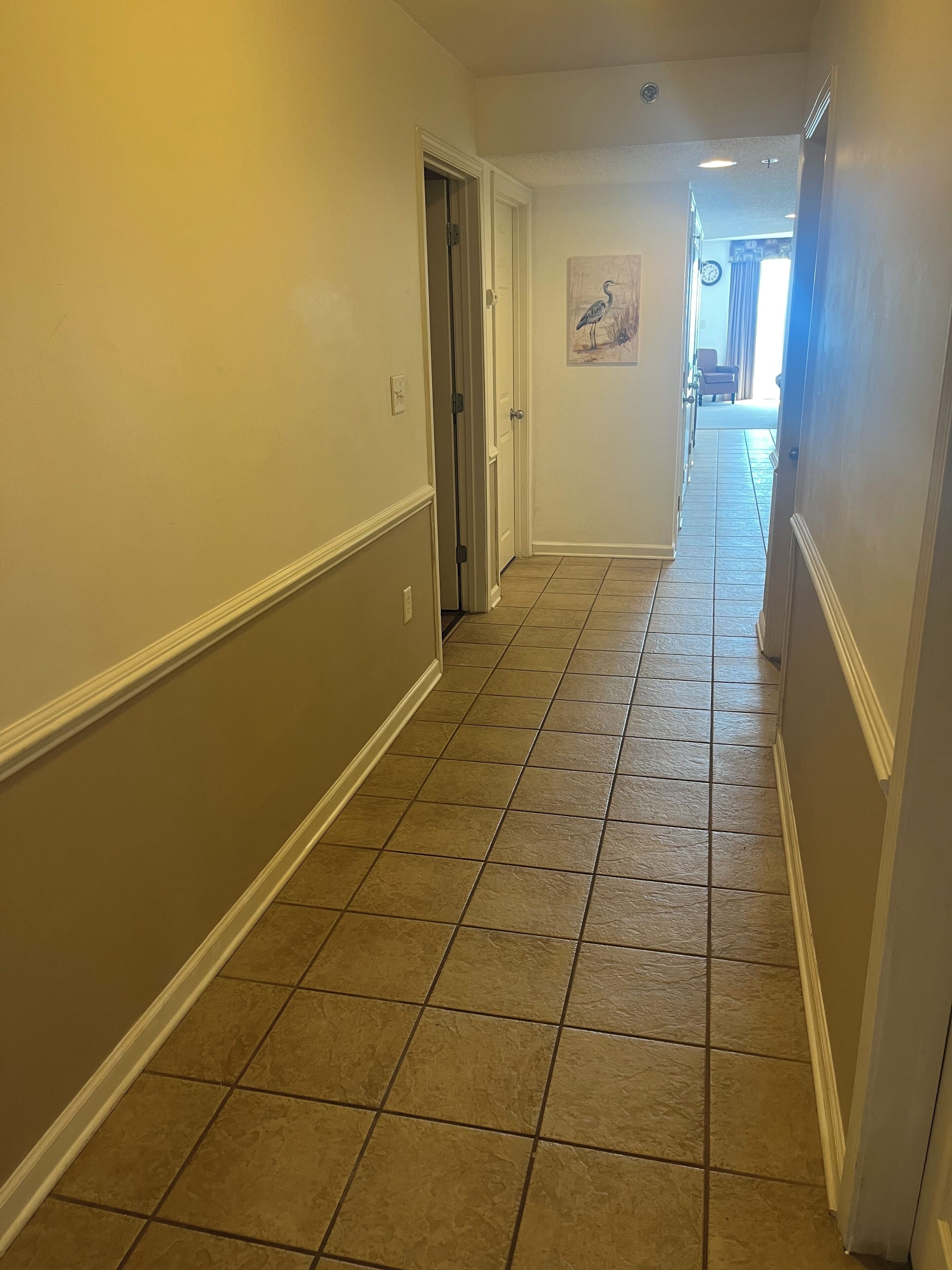 A hallway with beige walls and a tiled floor, leading to a room with a door.