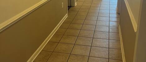 A hallway with beige walls and a tiled floor, leading to a room with a door.
