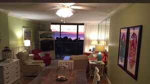 A living room with a wooden dining table, two sofas, a TV, and a balcony with a sunset view. - Dunes of Panama C302 — Sept 1-12 SPECIAL — with Deluxe Free BEACH CHAIR Rentals (Panama City)