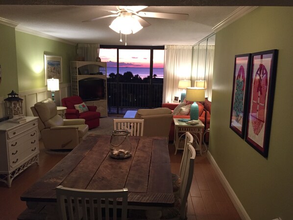 A living room with a wooden dining table, two sofas, a TV, and a balcony with a sunset view.