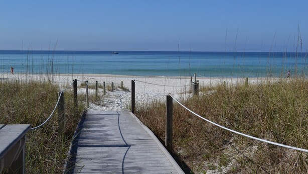 A wooden boardwalk leading to a sandy beach with clear blue water and a boat in the distance. - Dunes of Panama C302 — Sept 1-12 SPECIAL — with Deluxe Free BEACH CHAIR Rentals (Panama City)