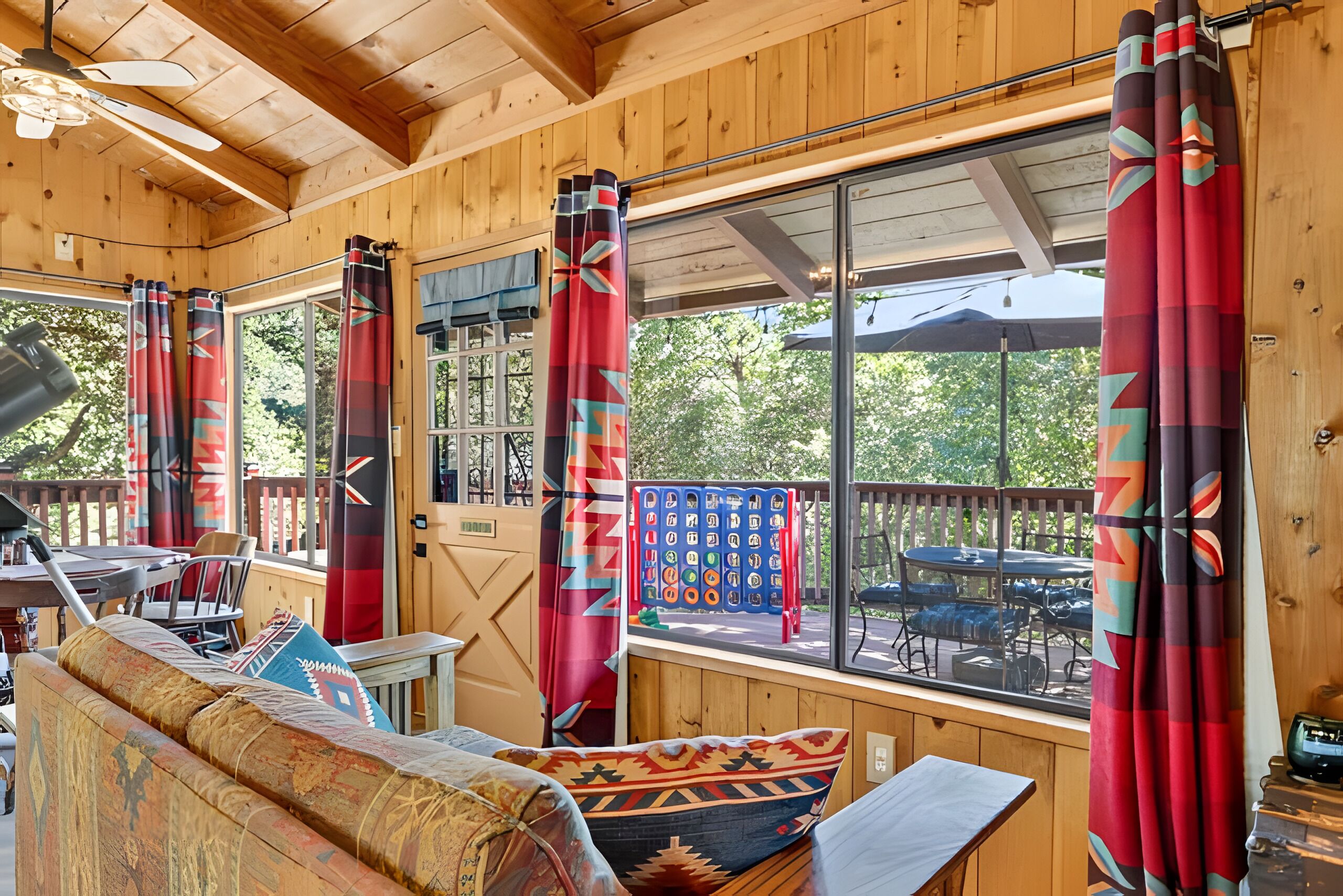 A cabin interior with a wooden ceiling, large windows with curtains, a sofa, and a table with chairs.