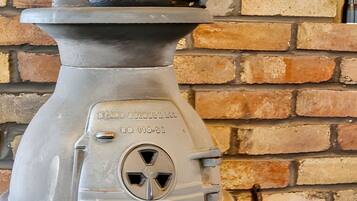 A vintage stove with a metal kettle on top, a brick wall background, and a clock on the wall.