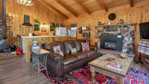 A cozy cabin interior with a wooden ceiling, a fireplace, a leather sofa, and a dining table.