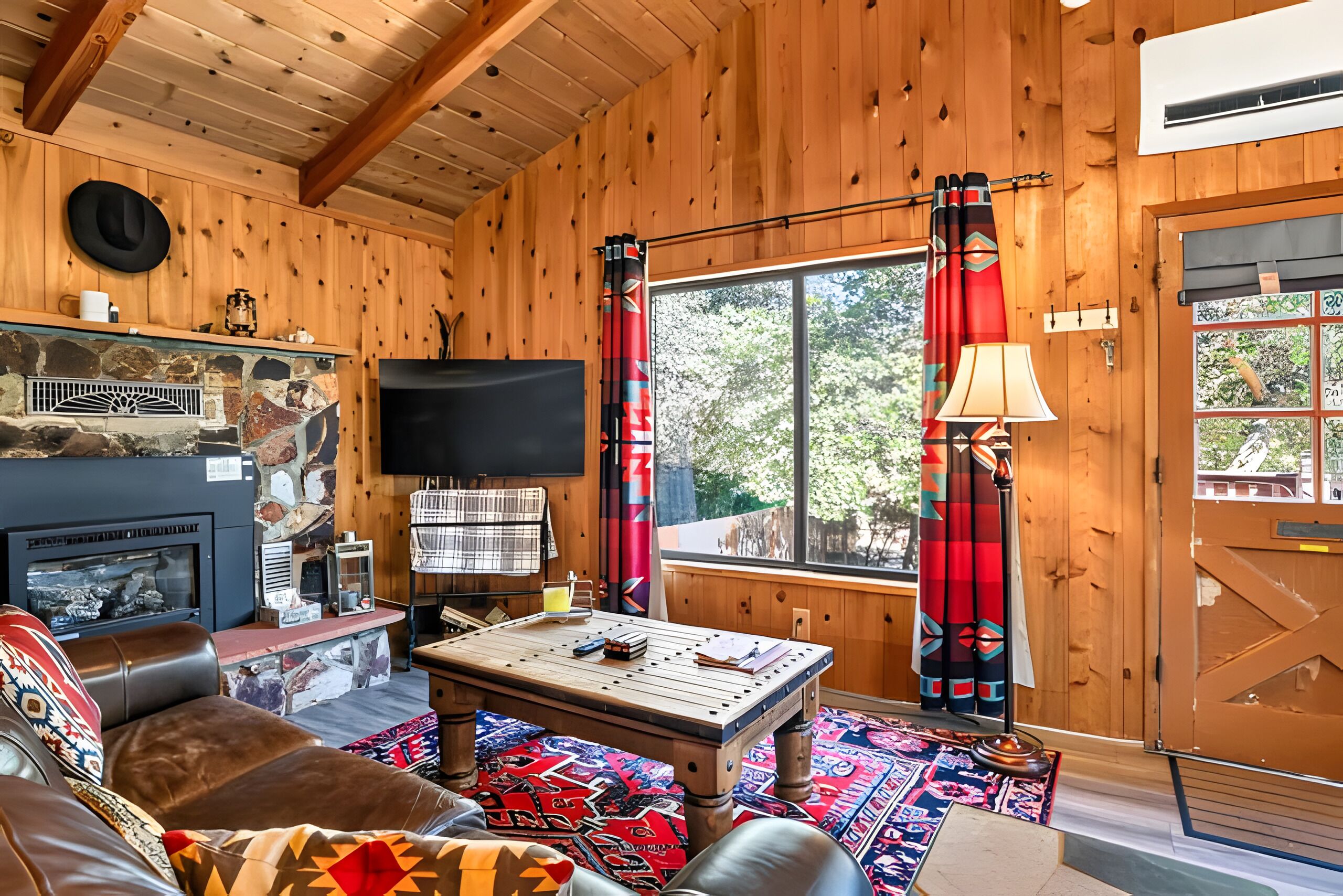 A cozy cabin interior with a fireplace, a television, a dining table, and a large window with curtains.