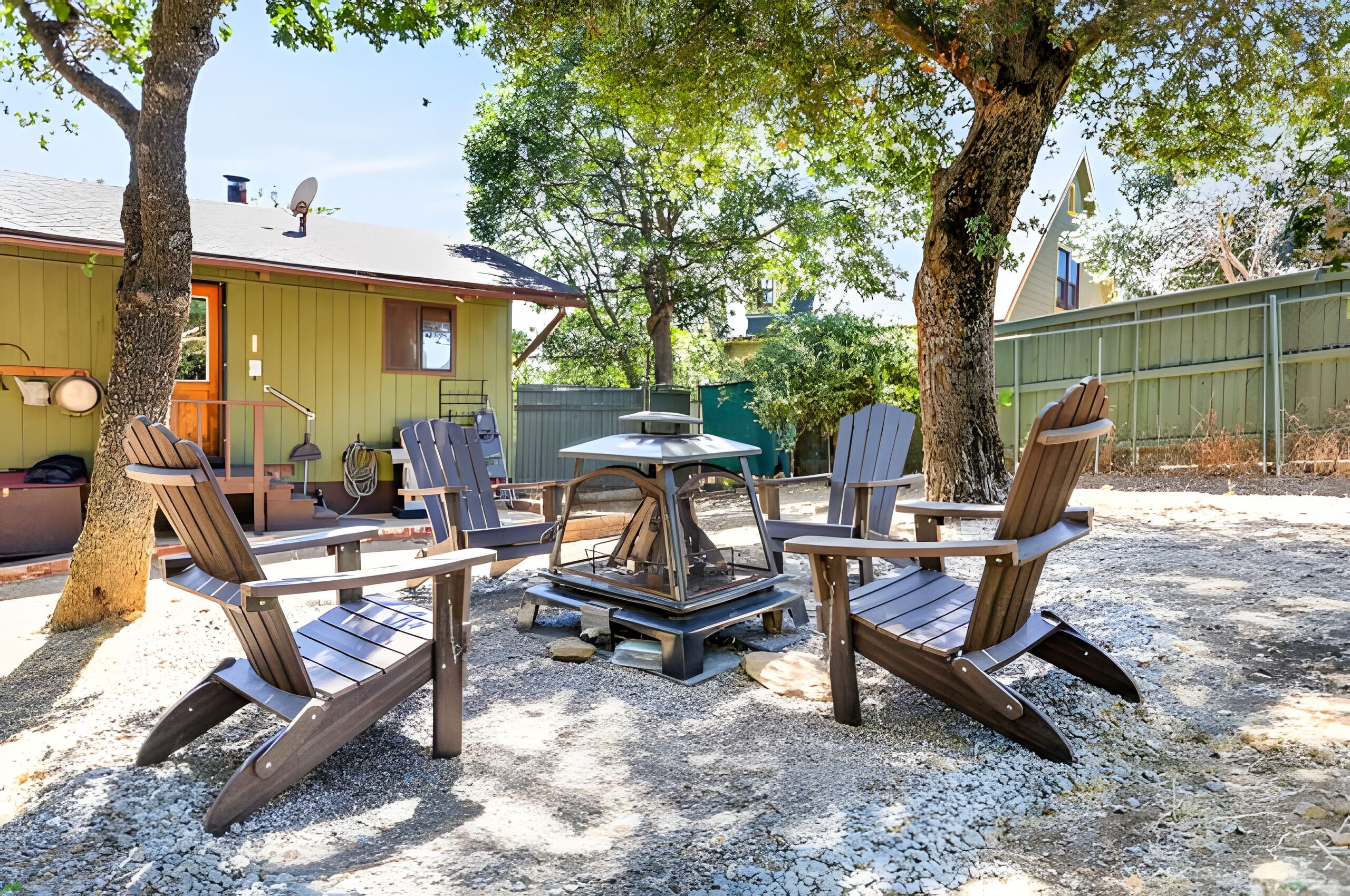 An outdoor seating area with Adirondack chairs and a central table, surrounded by trees and a building with a green exterior.