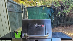 A stainless steel outdoor grill with a digital display, a side table, and a red bucket.