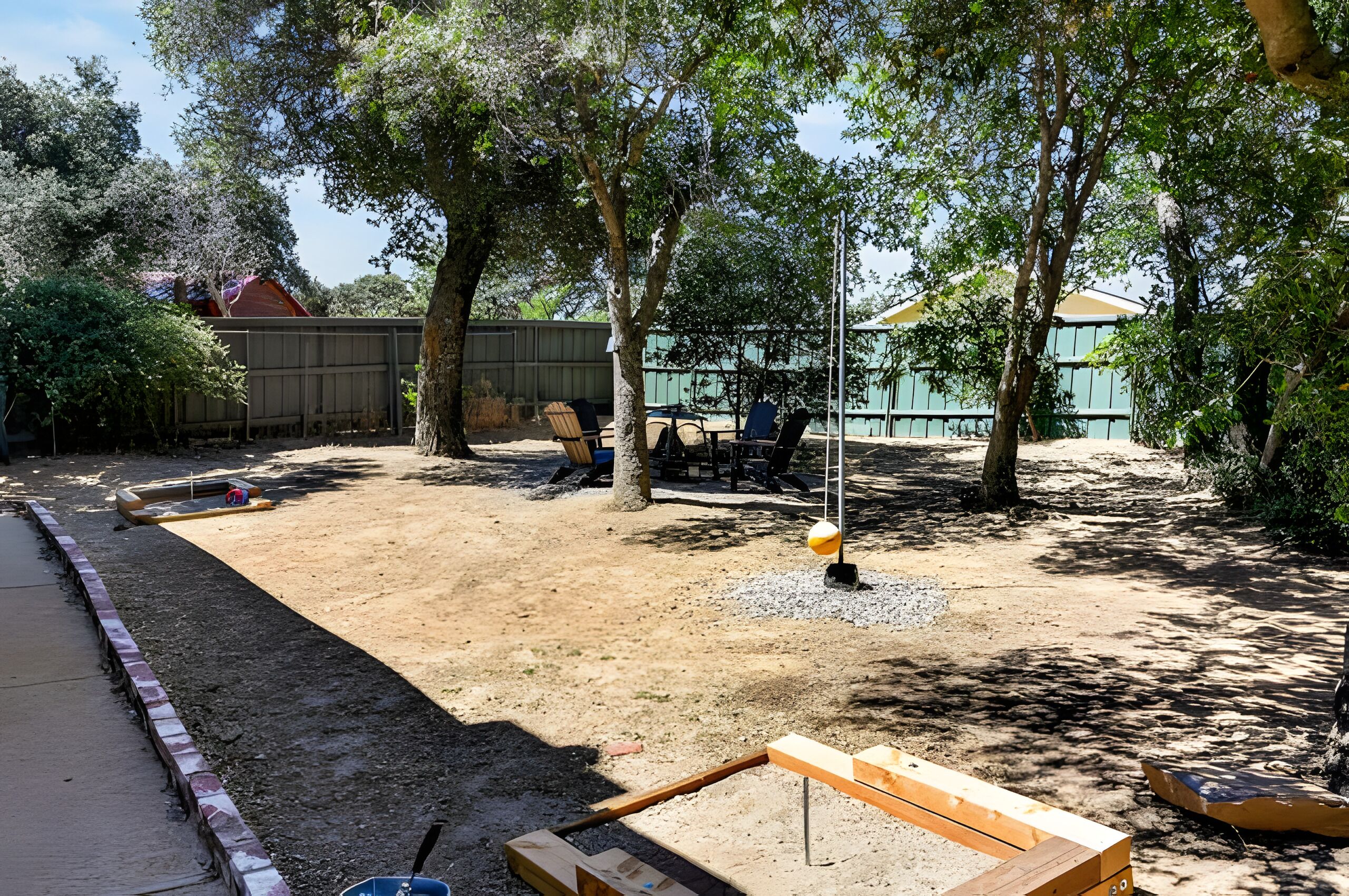 A backyard with a dirt area, a wooden deck, and a green fence.