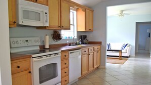 A kitchen with wooden cabinets, a microwave, stove, and dishwasher. There is a window and a door leading to a living area with a couch. - Spacious, light-filled mid-century home in Old Town (Bay Saint Louis)