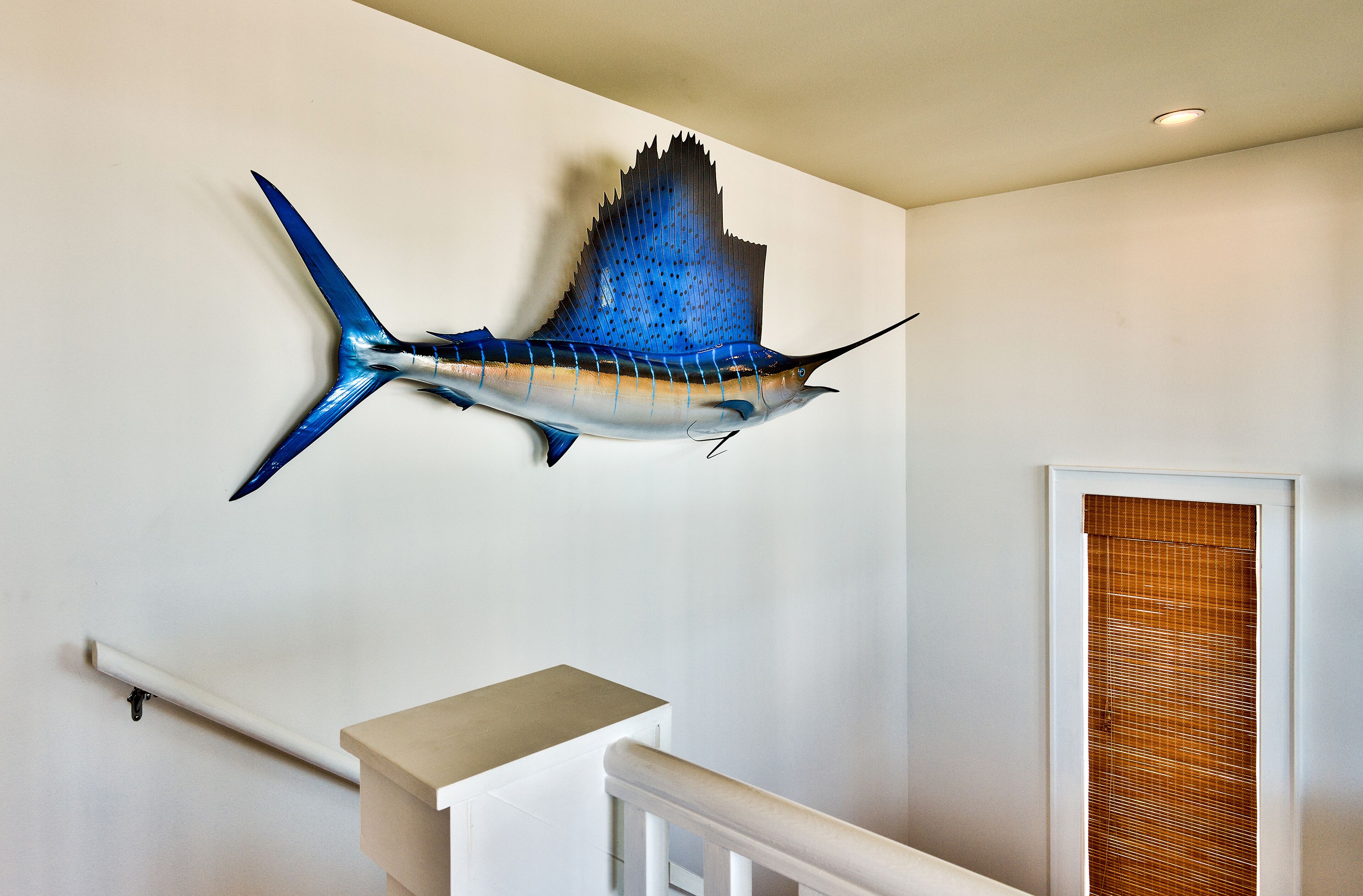 A wall-mounted sculpture of a sailfish in a hallway with a white railing and a door with a bamboo blind.