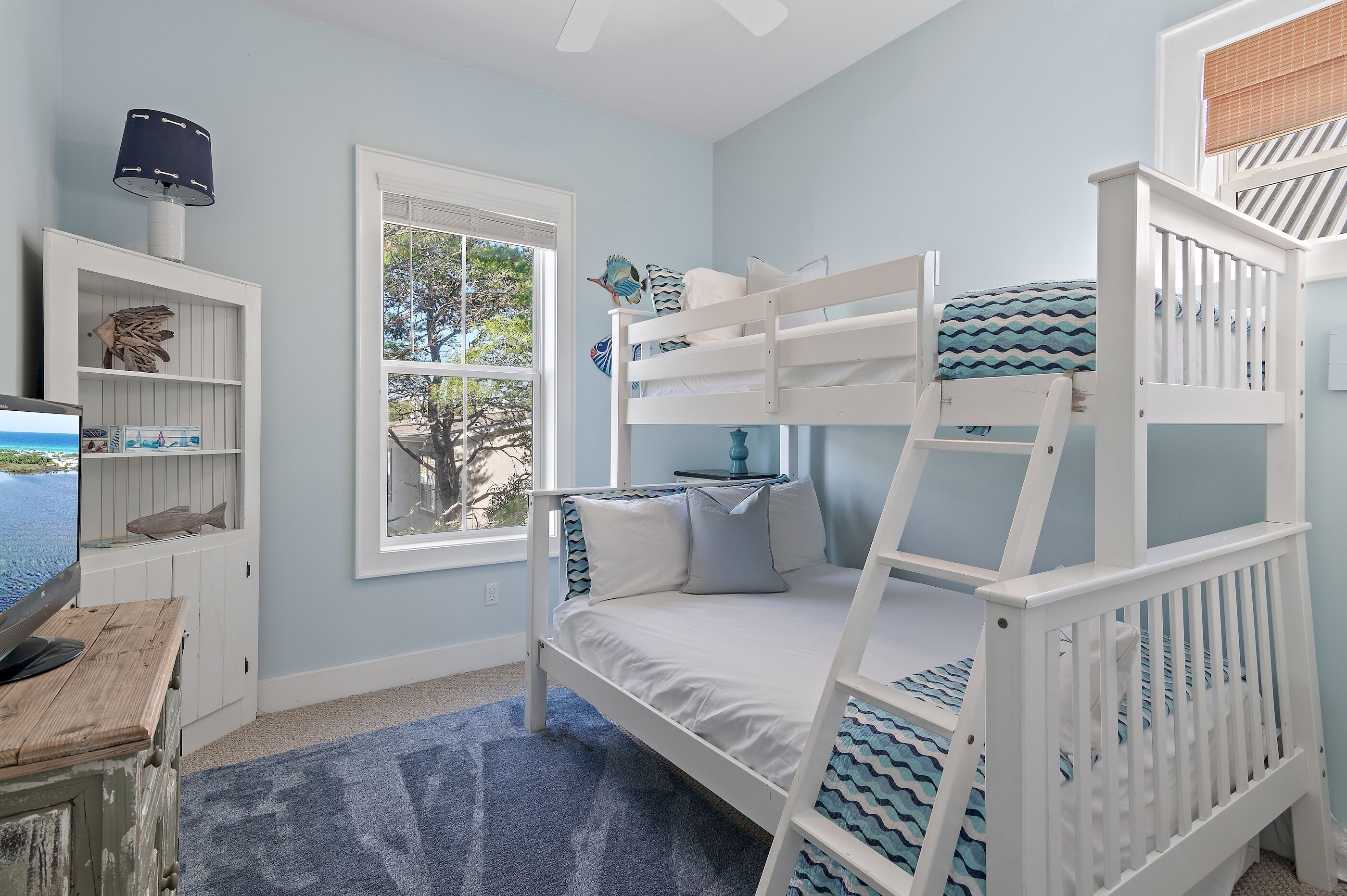 A child's bedroom with bunk beds, a TV, a bookshelf, and a window with a view of trees.