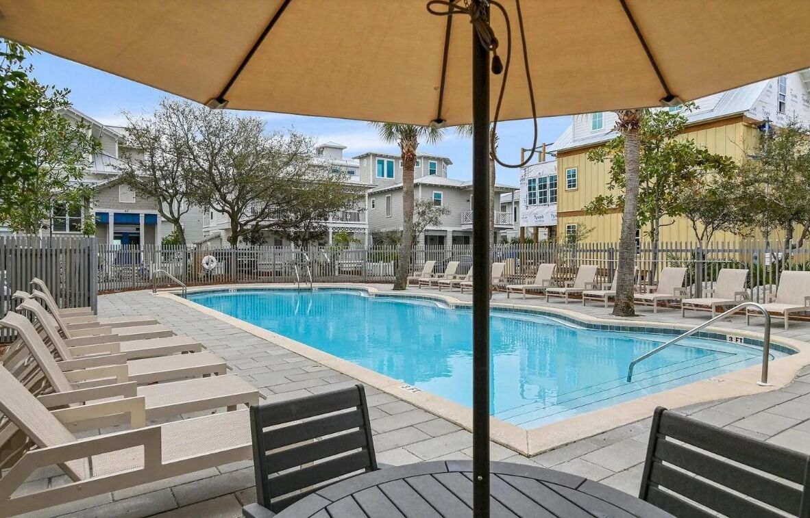 A swimming pool with lounge chairs and a table, surrounded by a fence and buildings.